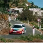Coche de rally Honda compitiendo en una curva cerrada en una carretera montañosa, con espectadores observando desde la ladera. Señales de tráfico indican límites de velocidad.