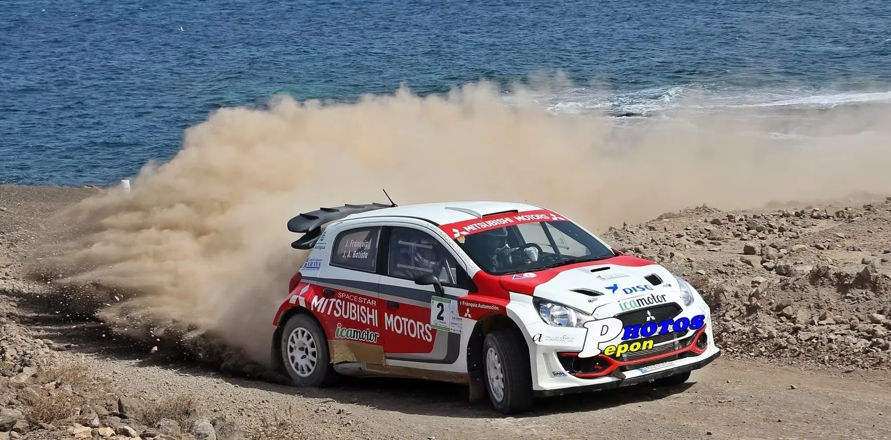 Coche de rally Mitsubishi en acción, levantando polvo en un camino costero, con el océano al fondo.